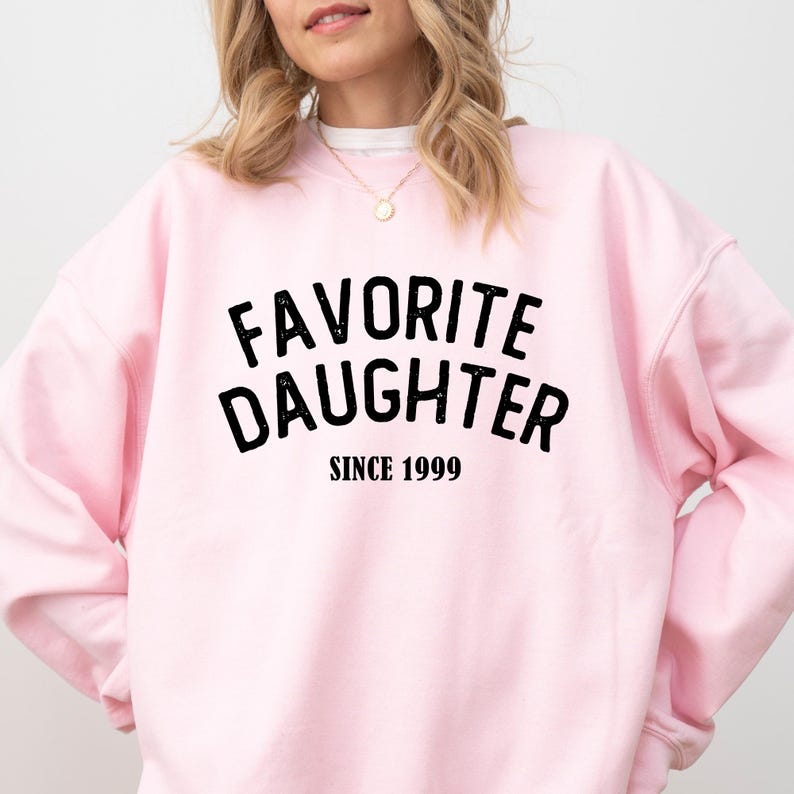 A woman wearing a pink sweatshirt with the text "Favored Daughter Since 1999" printed on it.