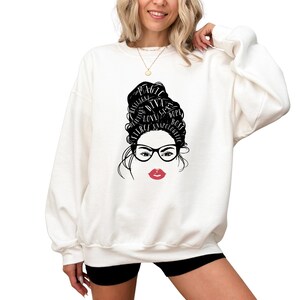 May include: A white crewneck sweatshirt featuring a graphic print of a stylized face with black glasses and red lips. The hair is depicted as a high bun containing the words Magic, Diva, Dope, Boss, Unapologetic, Fierce, Love, Sexy, and Beautiful.