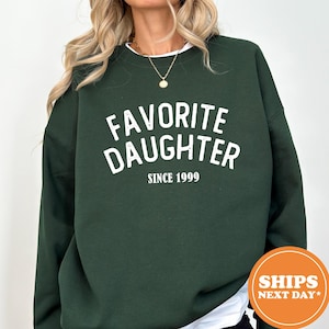 A person wearing a green sweatshirt with the text "Favored Daughter Since 1999" printed on it.