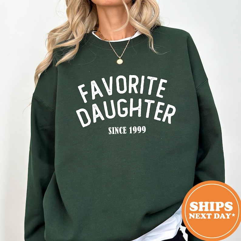 A person wearing a green sweatshirt with the text "Favored Daughter Since 1999" printed on it.