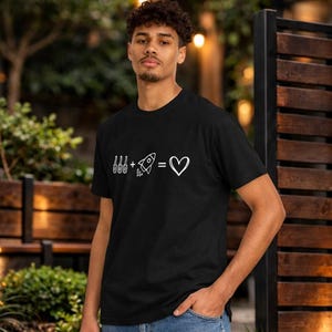 May include: Black t-shirt featuring a white graphic. The design shows three vertical lines, a rocket, an equals sign, and a heart symbol. The t-shirt is worn by a person.