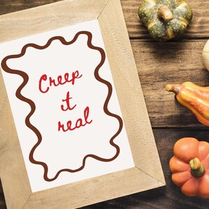 May include: A framed print with the words "Creep it real" in red script, surrounded by a brown wavy border. The frame is light brown. Decorative pumpkins in various colors are arranged on a wooden surface.