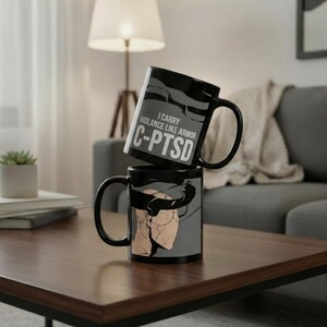 Könnte beinhalten: Zwei schwarze Keramikbecher, die auf einem Holztisch gestapelt sind. Der obere Becher hat den Text "I carry vigilance like armor C-PTSD". Der untere Becher zeigt eine Grafik eines Kopfes. Die Becher haben schwarze Griffe.