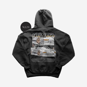 Op de afbeelding: Een antracietkleurige hoodie met een grafische print van een zilveren Honda NSX. De grafiek bevat de tekst "HONDA NSX" in een gouden omtrek. De tekst "1991 HONDA NSX" is onder de grafiek gedrukt.