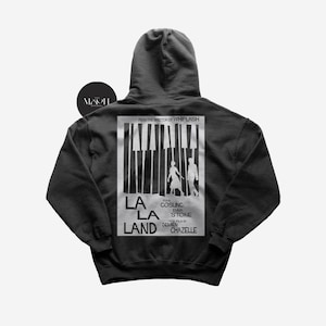 May include: Black hooded sweatshirt with a white print of the La La Land movie poster on the back. The poster features the film's title, the names of the actors Ryan Gosling and Emma Stone, and the director Damien Chazelle.