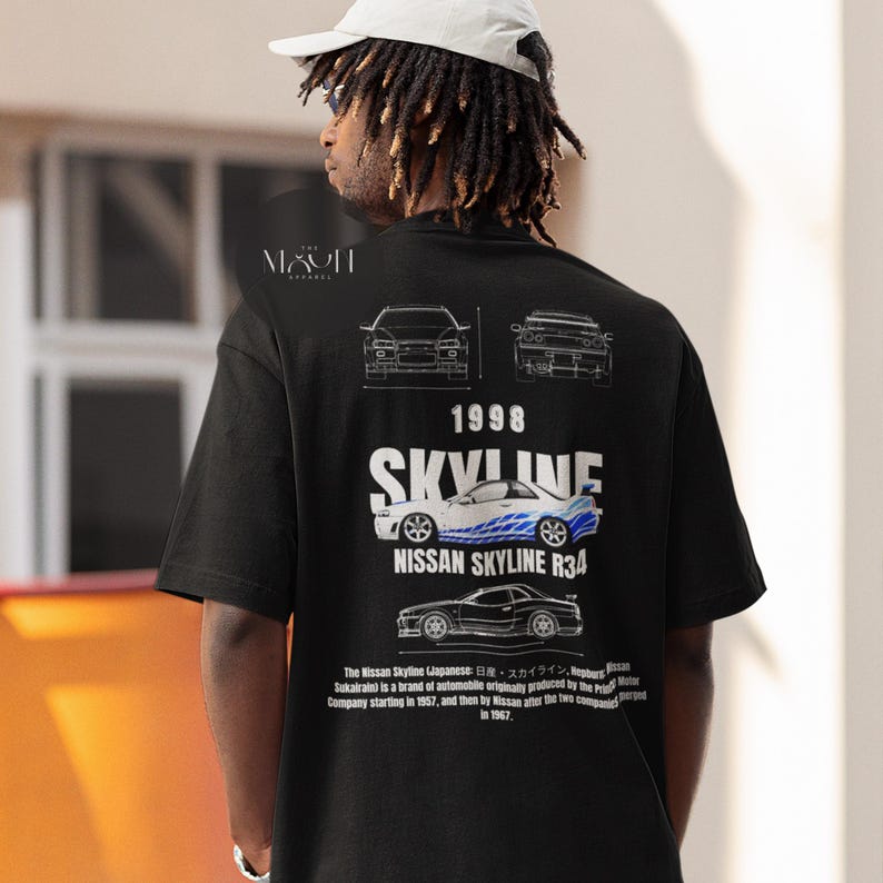 May include: Black t-shirt with a white graphic print of a blue Nissan Skyline R34 car. The graphic includes the text "1998 SKYLINE NISSAN SKYLINE R34". The text below the car reads "The Nissan Skyline (Japanese: 日産・スカイライン, Hepburn: Sukairain) is a brand of automobile originally produced by the Prince Motor Company starting in 1957, and then by Nissan after the two companies merged in 1967."