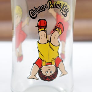 May include: Clear glass drinking glass featuring a Cabbage Patch Kids design. The glass has a cartoon image of a child wearing yellow overalls, red shirt, and red shoes. The words "Cabbage Patch Kids" are printed at the top.