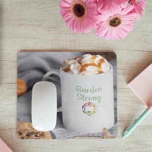 Puede incluir: Una taza de cerámica blanca con un diseño marrón y blanco. La taza está llena de una bebida cremosa cubierta con caramelo y crema batida. La taza tiene el texto "Garden Strong" y un logotipo verde con un círculo de hojas de colores. La taza está sobre una alfombrilla de ratón gris y marrón con un ratón de ordenador al lado.