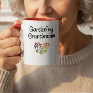 Peut inclure: Mug en céramique blanche avec intérieur et anse rouges. Le mug porte l'inscription "Gardening Grandmama" en écriture noire, ainsi qu'un motif en forme de cœur rempli de légumes et de fleurs colorés. Le mug est tenu par une personne.