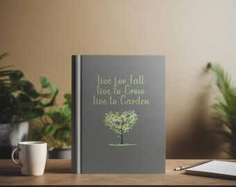 Può includere: Un diario grigio con il testo "live for Fall live to Grow live to Garden" e un'illustrazione di un albero a forma di cuore. Una tazza bianca, una penna e un blocco note sono su una superficie di legno, con piante sullo sfondo.