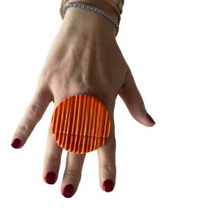 Könnte beinhalten: Ein großer, runder Ring mit geriffelter Textur in leuchtend orange. Der Ring wird an einem Finger getragen.