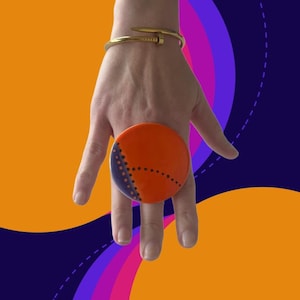 Puede incluir: Un anillo circular grande, de color naranja, con una sección azul oscuro y un diseño punteado. El anillo se lleva en una mano, con una pulsera dorada en la muñeca. El fondo es una mezcla de naranja, morado y azul.