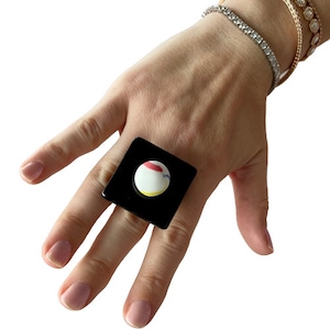 Può includere: Un anello quadrato nero con un cerchio bianco al centro. Il cerchio ha un design giallo, rosso e blu.