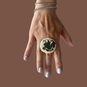 Op de afbeelding: Een witte ring met een zwart abstract ontwerp. De ring wordt gedragen op een vinger met lichtblauwe nagellak.