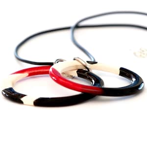 Op de afbeelding: Een ketting met een zwart koord en een hanger gemaakt van twee in elkaar grijpende ringen. De ringen zijn zwart, wit en rood geverfd.