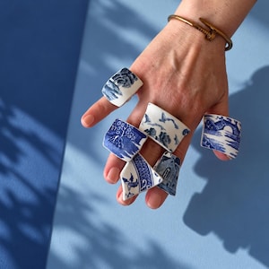 May include: A collection of five rings featuring curved, white ceramic shards decorated with blue transferware patterns depicting landscapes, architectural structures, and floral motifs. Each ring displays a unique fragment of blue and white pottery.