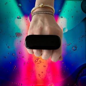 Puede incluir: Un objeto negro rectangular, sostenido en una mano adornada con pulseras doradas y plateadas. El objeto tiene una superficie lisa y brillante. El fondo presenta un diseño abstracto colorido con tonos de azul, rojo y verde.