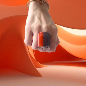 May include: A bold, rectangular ring with a clear base and a colourful top. The top section is split into two vertical segments: one bright orange and the other dark grey. The ring is worn on a hand, with a gold bracelet visible.
