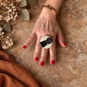 May include: A circular ring featuring a cream-colored base with a black diagonal stripe and a small black spiral accent. The ring is worn alongside a gold-toned metal cuff bracelet with a nail-inspired design on the wrist.
