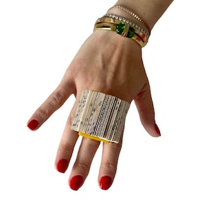 May include: A yellow ring made of pages from a book. The ring is worn on a finger with red nail polish.