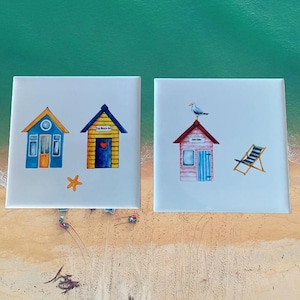 Puede incluir: Dos baldosas de cerámica cuadradas con ilustraciones de temática playera. Una baldosa presenta dos cabañas de playa coloridas, una estrella de mar y el texto "The Beach Hut". La otra baldosa muestra una cabaña de playa, una gaviota y una tumbona.
