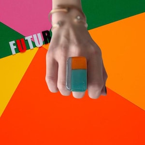 May include: A rectangular glass ring featuring a colour-blocked design with a horizontal orange section at the top and a larger teal section below, set against a grey vertical strip. The word FUTUR is visible in the background in white and red letters.
