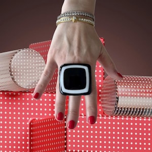 Op de afbeelding: Een grote, vierkante, zwart-witte glazen ring met een witte rand. De ring wordt gedragen aan de wijsvinger van een hand.