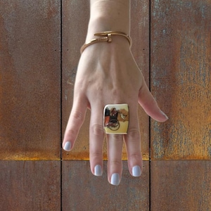 May include: A square ring with a painted scene featuring figures and a carriage. The ring is set on a hand with light blue nail polish, and a gold-colored bracelet is on the wrist. The background is a rusty brown.