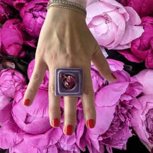 Op de afbeelding: Een vierkante ring van paarse kleur met een gedroogde bloem erin. De ring zit aan een vinger, en de achtergrond is een boeket roze pioenen.