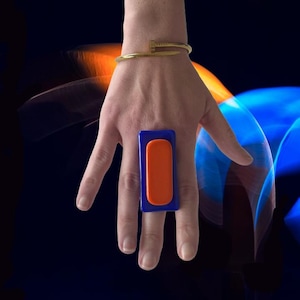 Pode incluir: Um anel retangular arrojado com um centro laranja vibrante emoldurado por uma moldura azul profunda. O anel é uma peça de joalheria marcante. Uma pulseira dourada também é visível.