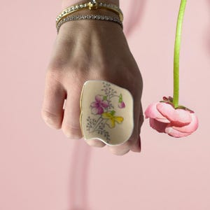 Könnte beinhalten: Ein Ring aus einem Stück Vintage-Porzellan mit einem floralen Design in Rosa, Gelb und Grün. Der Ring wird an einer Hand mit einem Armband getragen.