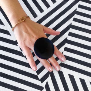 May include: A large, circular, dark blue ring with a smooth, reflective surface worn on the middle finger, paired with a thin, gold-toned metal nail-shaped bangle bracelet featuring a small screw-like detail on the end of the band.