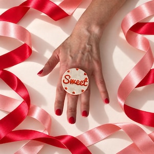 May include: A circular ring featuring a white surface with the word Sweet written in orange script, accented by small orange hearts. The ring is surrounded by flowing red and pink satin ribbons on a light background, creating a festive display.