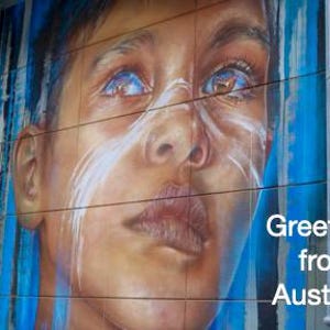 Pode incluir: Um mural com um retrato detalhado de uma pessoa com olhos azuis e marcas brancas no rosto. A obra de arte est&aacute; em uma parede de azulejos com listras verticais azuis e brancas. O texto diz "Greetings from Australia."