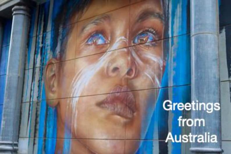 Pode incluir: Um mural com um retrato detalhado de uma pessoa com olhos azuis e marcas brancas no rosto. A obra de arte est&aacute; em uma parede de azulejos com listras verticais azuis e brancas. O texto diz "Greetings from Australia."