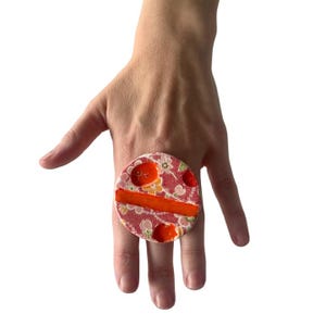 May include: A large, round ring with a red floral fabric design and a bright orange accent. The ring is set on a hand, showcasing its size and color contrast. The ring's design features a mix of red, orange, and white floral patterns.