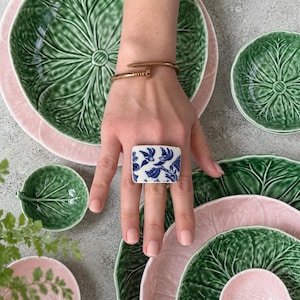 May include: A gold-toned nail-shaped bangle bracelet and a rectangular ceramic ring featuring a blue and white bird pattern. The ring is worn on a finger, showcasing a detailed illustration of birds perched on branches against a white background.