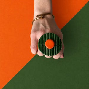 May include: A gold-toned metal nail-shaped bracelet worn on the wrist, paired with a circular, dark green, vertically ribbed object featuring a smooth, bright orange, raised circular center piece held in the palm of the hand.