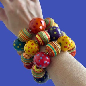 Könnte beinhalten: Ein farbenfrohes Perlenarmband mit großen, runden Perlen. Die Perlen weisen eine Vielzahl von Mustern auf, darunter Punkte und Streifen in Rot-, Gelb-, Orange-, Grün- und Blautönen. Das Armband wird am Handgelenk getragen.