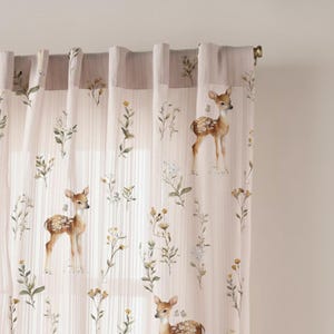 May include: Light pink curtains with a repeating pattern of deer and wildflowers. The deer are brown with white spots, and the flowers are yellow, white, and green. The curtains are hanging from a gold rod.