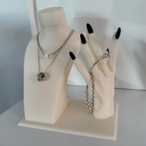 May include: A cream-colored jewelry display stand featuring a bust and a hand. The bust holds two silver necklaces with sparkling pendants. The hand displays a silver bracelet and a chain bracelet. The nails are painted black.