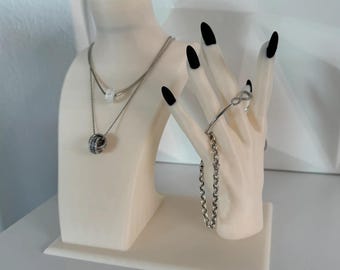 Op de afbeelding: Een crèmekleurige sieradenstandaard met een buste en een hand. De buste houdt twee zilveren kettingen met fonkelende hangers vast. De hand toont een zilveren armband en een kettingarmband. De nagels zijn zwart gelakt.