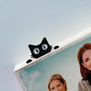 May include: A black cat-shaped bookmark peeks over the edge of a book. The bookmark has large, white eyes and small whiskers. The book has a picture of two women and a baby.