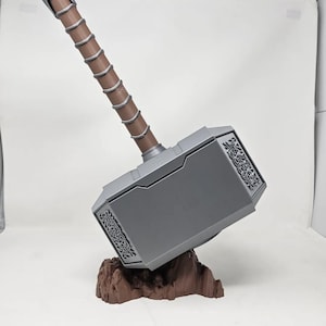 May include: A silver-colored Mjolnir hammer with a brown handle, resting on a brown rock-like base. The hammer has intricate details and a design on the side.
