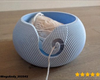 May include: A light blue yarn bowl with a spiral design and a light-colored yarn ball inside. The bowl has a textured, ribbed exterior and a smooth, blue interior. The yarn thread exits through a spiral opening.