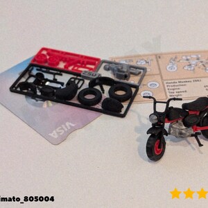 Op de afbeelding: Een miniatuur zwart en rood motorfietsmodel met rode wielen en een zwart zadel. Het model staat naast niet-gemonteerde onderdelen op een zwart plastic frame en een kaart met het VISA-logo.