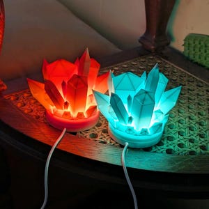 Könnte beinhalten: Zwei kristallförmige LED-Lampen, eine rote und eine türkisfarbene, stehen nebeneinander. Jede Lampe hat einen weißen Sockel und eine Ansammlung geometrischer Kristallformen. Die Lampen sind mit weißen Kabeln angeschlossen und stehen auf einem Holztisch.