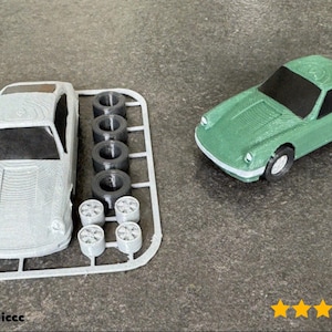 Op de afbeelding: Een groene 3D-geprinte modelauto naast een gedemonteerde carrosserie en wielen. De geassembleerde auto is groen met zwarte ramen en witte wielen. De niet-geassembleerde auto is grijs. De wielen zijn zwart en zilver.