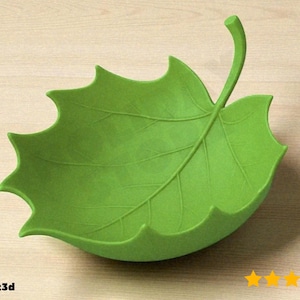 May include: A bright green, leaf-shaped bowl with detailed vein patterns. The bowl has a curved stem and scalloped edges, resembling a large leaf. The bowl is made of a solid material and sits on a light wood-grain surface.