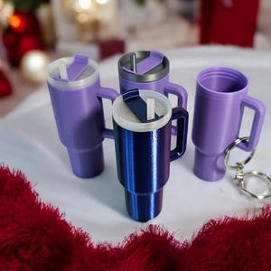 May include: Four miniature tumblers with lids and straws. Two are purple, one is blue, and one is silver. The tumblers are on a white surface with a red fuzzy blanket in the foreground.